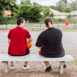 Two young men sitting on a bench near a basketball court