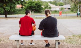 Two young men sitting on a bench near a basketball court