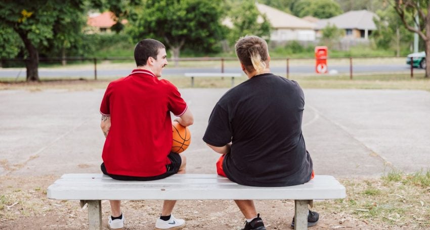 Two young men sitting on a bench near a basketball court