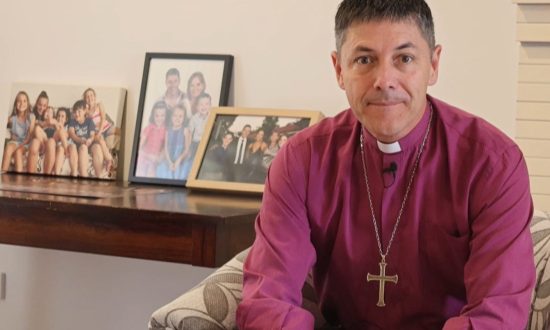 Archbishop in purple shirt and collar sitting in armchair with framed family images behind him