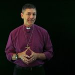 Archbishop Jeremy Greaves wearing a purple shirt and standing against a black background