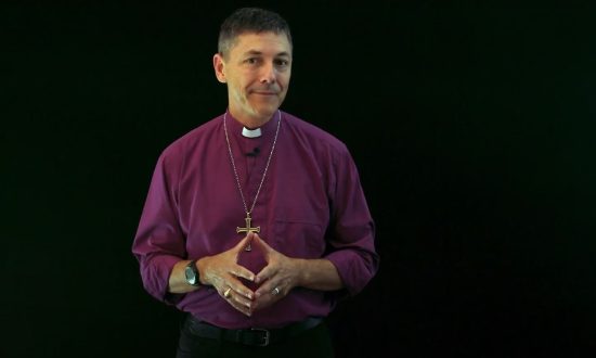 Archbishop Jeremy Greaves wearing a purple shirt and standing against a black background