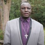 Bishop Daniel Abot standing under gum trees near a wooden cross