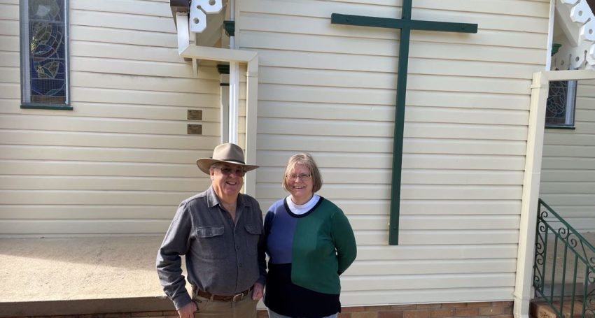 A man wearing an akubra and a woman wearing jeans -- both priests -- standing outside a rural church made of timber