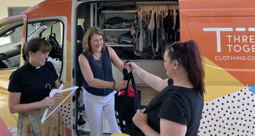 A woman priest and Anglicare Southern Queensland staff are assisting community members with new clothing in front of a Thread Together van