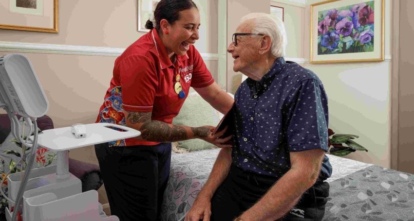 Nurse wearing red shirt taking the blood pressure of an older man in his room