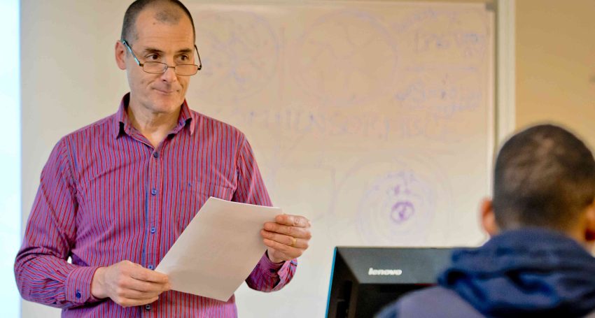 Man wearing purple shirt lecturing in front of a whiteboard