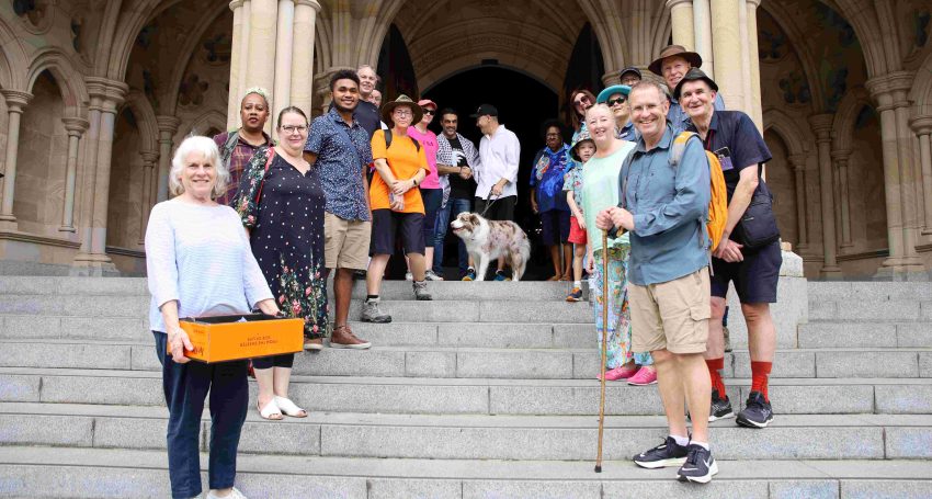 A large group of diverse people and a dog on cathedral steps during the middle of the day, with the people wearing walking gear
