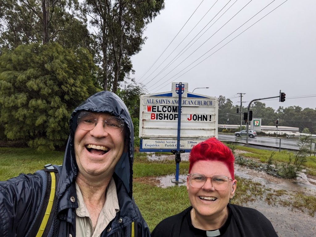 A bishop and a priest walking in the rain outside a church sign 