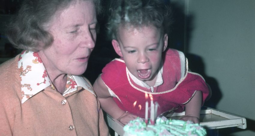 Toddler in the 1970s about to blow out birthday cake candles with grandmother sitting next to him