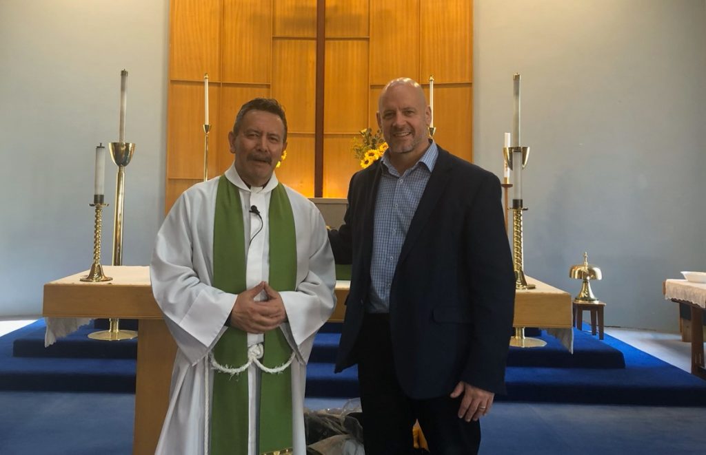 A priest in white robes and a man in a suit standing in front of a church altar