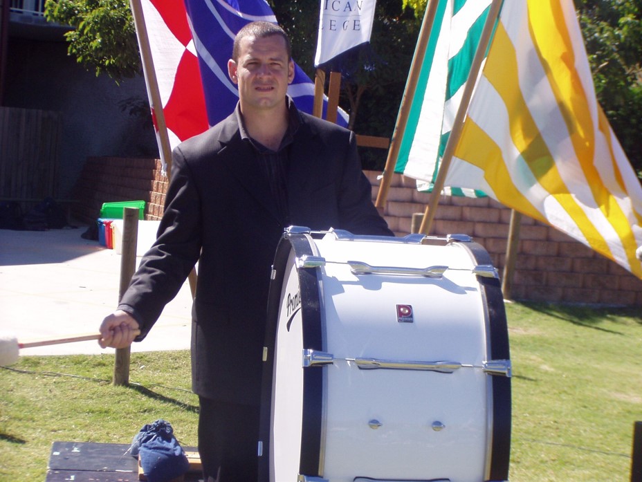 Young man in black beating a large white drum with flags behind him at a school