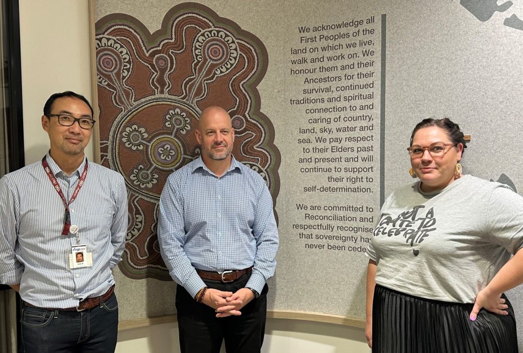 Three people standing in front of an Acknowledgement of Country artwork and text on an office wall 