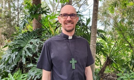 Male priest in black shirt and collar wearing a large green cross standing in front of bushes and trees