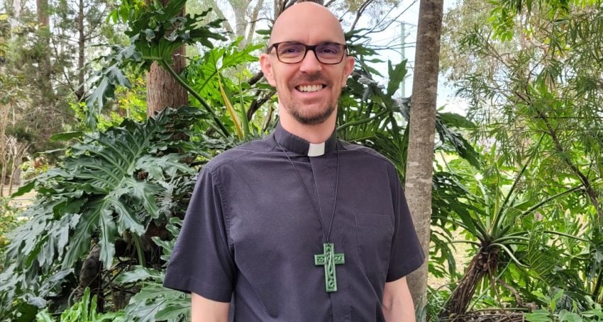 Male priest in black shirt and collar wearing a large green cross standing in front of bushes and trees