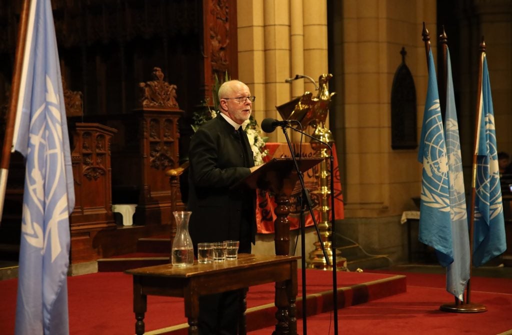 Priest in black garb and collar speaking into a microphone in a cathedral 