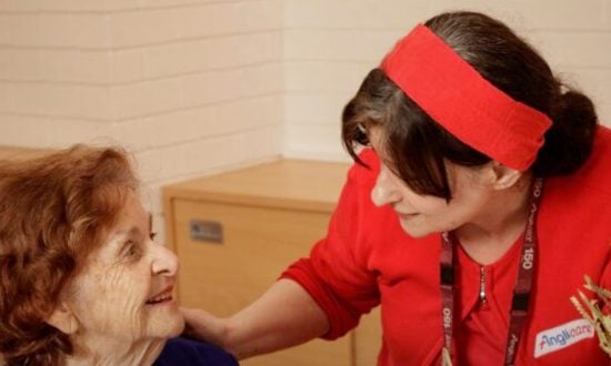 Nurse wearing red assisting older woman wearing blue and white in an aged care home