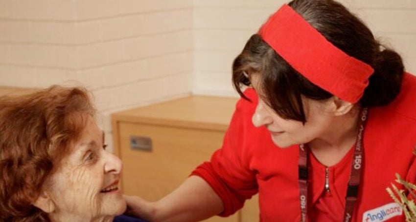 Nurse wearing red assisting older woman wearing blue and white in an aged care home