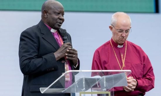 Two male bishops wearing purple shirts speaking at a podium