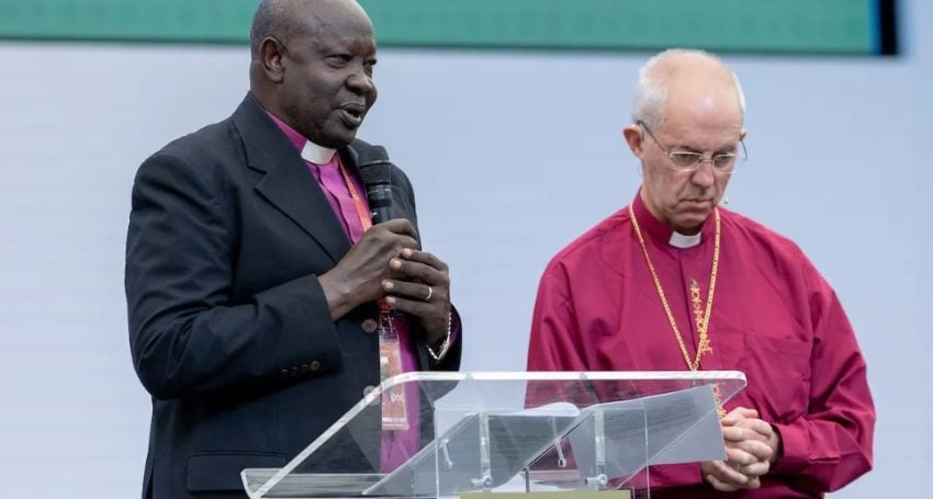 Two male bishops wearing purple shirts speaking at a podium