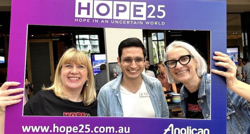 Two women preists and a man priest behind a photo board smiling