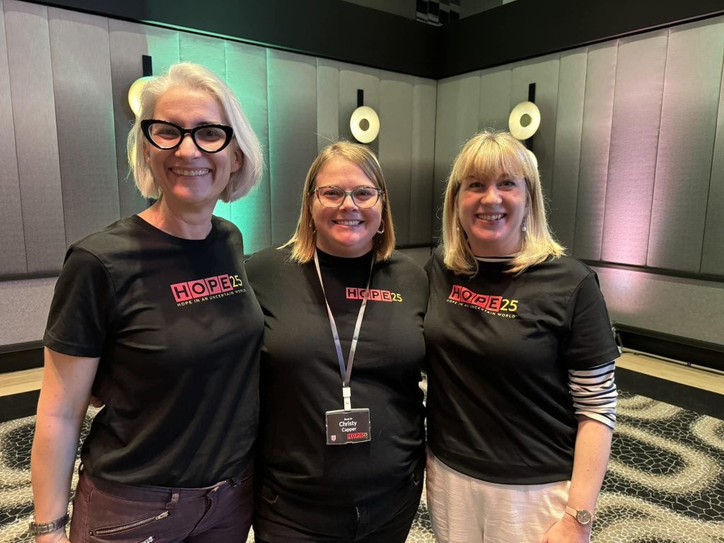 Three women priests wearing black t-shirts standing behind a photoboard smiling 