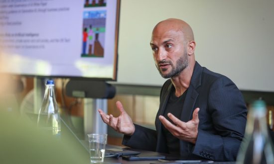 Man wearing black presenting with a big screen in the background