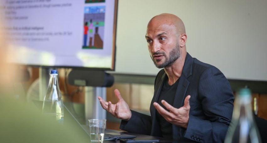 Man wearing black presenting with a big screen in the background