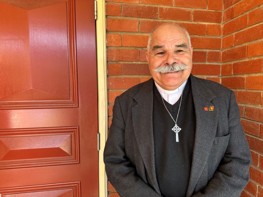 Aboriginal priest smiling in front of a red brick wall 