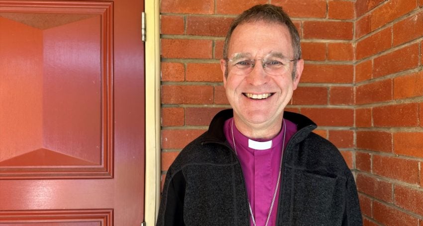 Bishop in purple shirt and black jacket smiling while standing next to a red brick wall