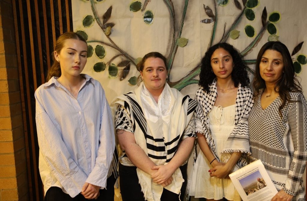 Four people of different faiths gathered in front of a painted olive tree, including one man and three women