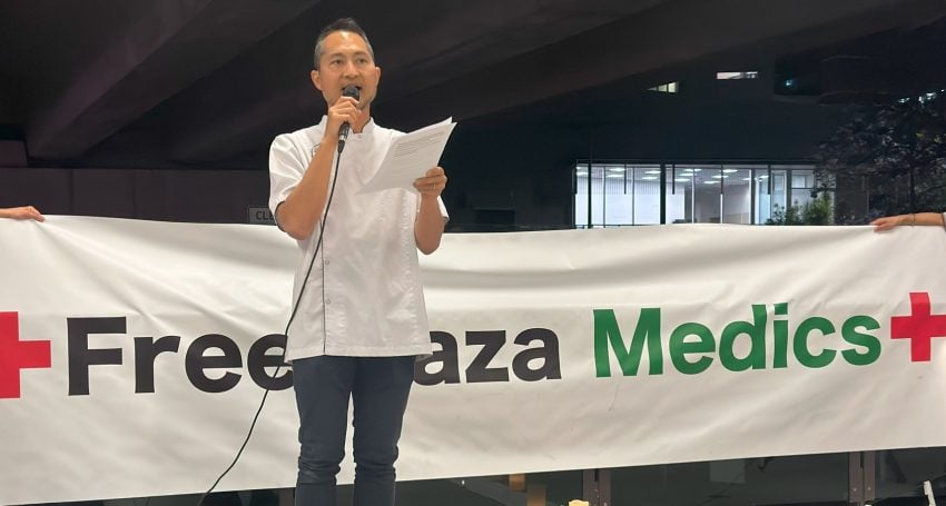 Pharmacist standing in front of a "Free Gaza Medics" sign giving a speech