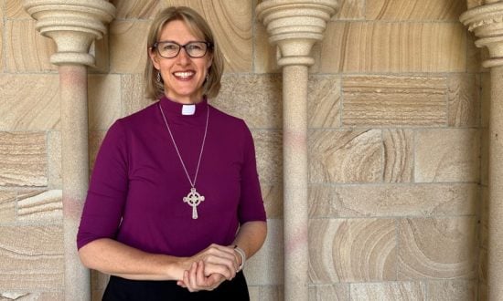 Woman bishop in purple shirt and collar in front of sandstone Cathedral wall