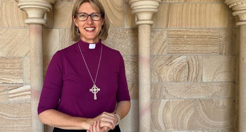 Woman bishop in purple shirt and collar in front of sandstone Cathedral wall