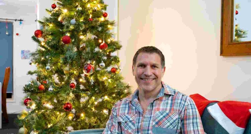 Man in plaid shirt sitting next to a Christmas tree