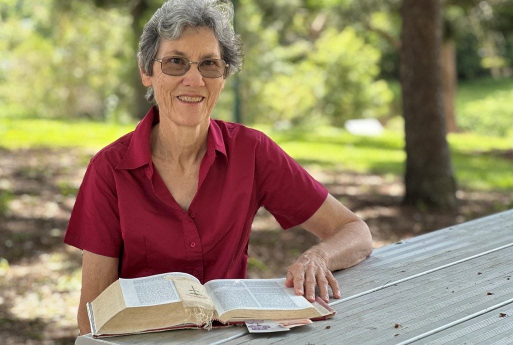 Woman in maroon shirt reading the bible at a park bench 