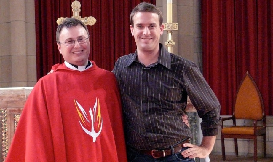 A priest in robes with a man hugging and smiling at the camera