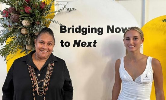 Two women stand in front of a sign that reads "Bridging Now to Next"