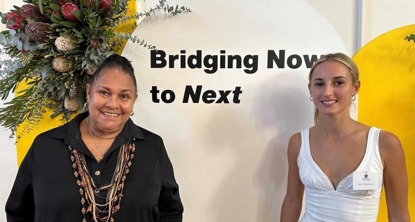 Two women stand in front of a sign that reads "Bridging Now to Next"