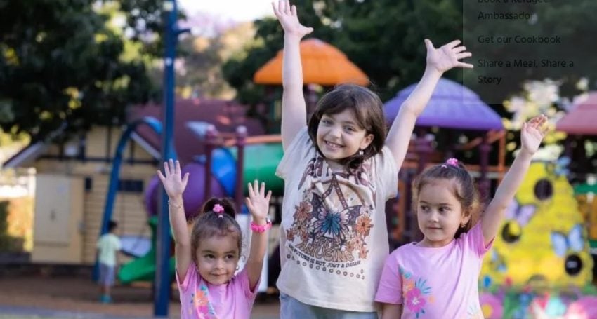 Three girls with their arms in the air smiling near a playground