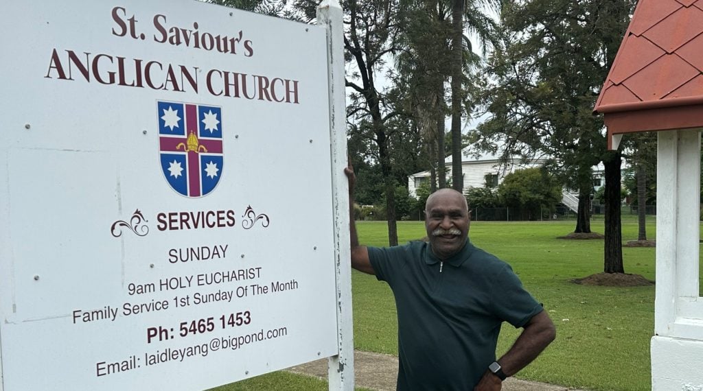 Torres Strait Islander elder leaning against a church sign 