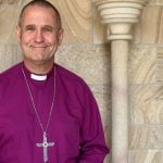 Bishop in purple shirt and collar and pectoral cross outside a cathedral entrance