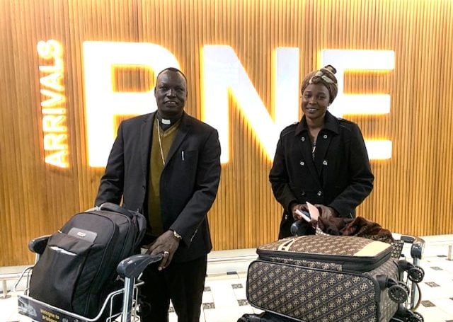 Two people pose for a photograph at an airport with luggage on trolleys
