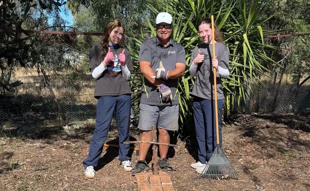 Two students and a teacher with garden tools in a rural backyard
