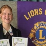 A female student stands in front of a Lions Club poster while displaying two award certificates