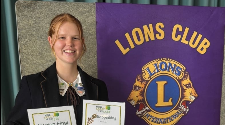 A female student stands in front of a Lions Club poster while displaying two award certificates