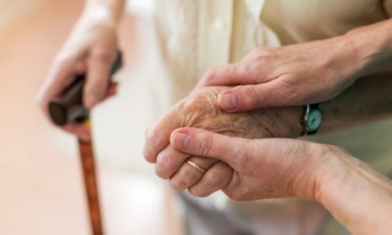 A carer's hands assist an elderly patient with a walking stick