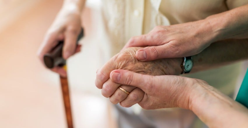 A carer's hands assist an elderly patient with a walking stick