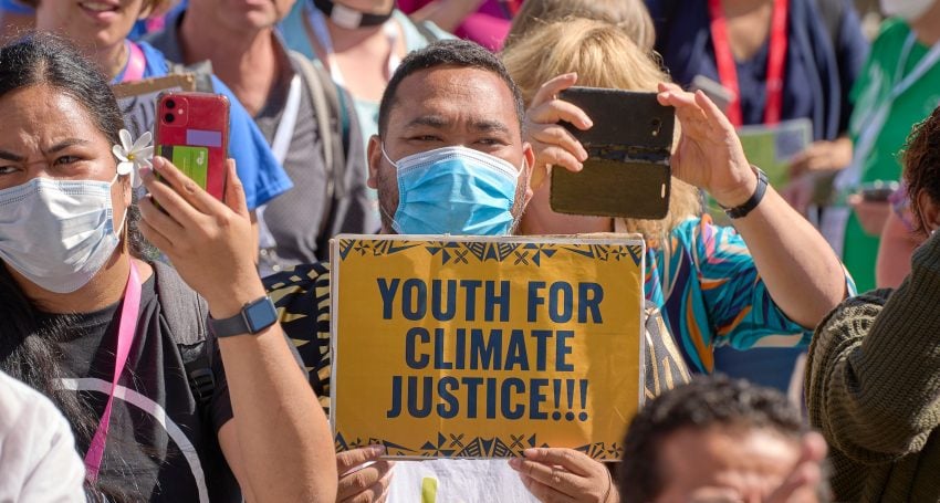 A group of climate protestors are gathered outside with one man holding a sign that reads "Youth for Climate Justice!"